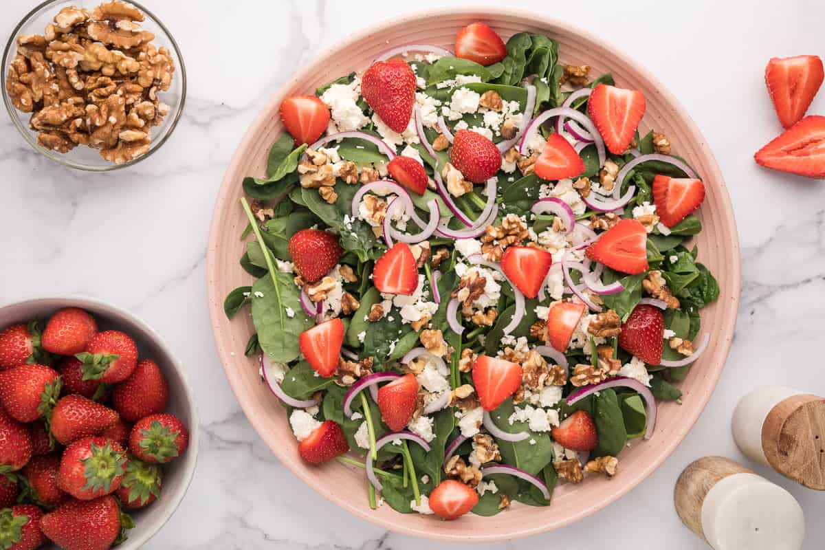 An undressed spinach strawberry salad on a pink plate with salad ingredients in bowls around it.