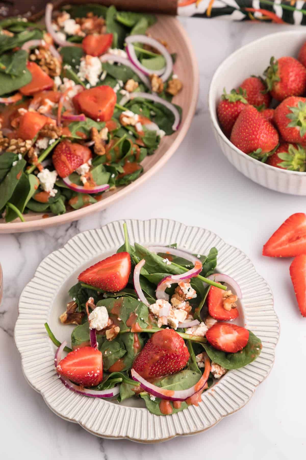 A serving on strawberry salad on a small white plate with a bigger plate of more salad and a bowl of strawberries in the background.