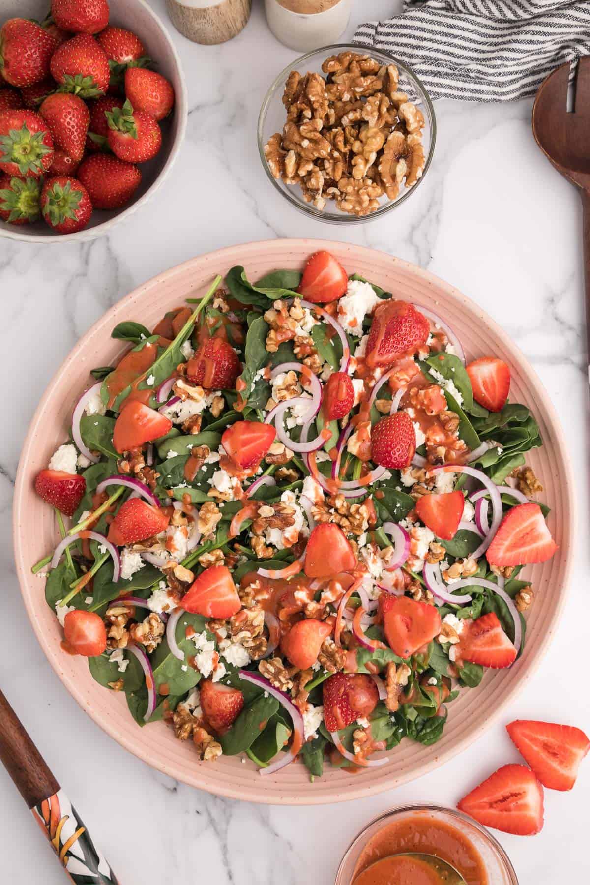 Overhead of a strawberry spinach salad with strawberry dressing on a large pink plate with salad ingredients around it.